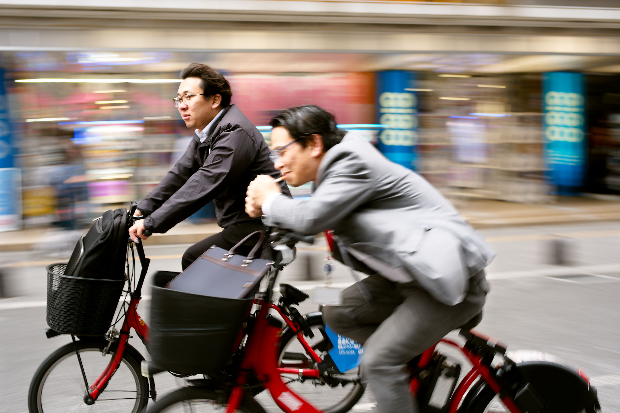 two japanese salarymen riding bicycles. one is leaned forward trying to light a cigarette.